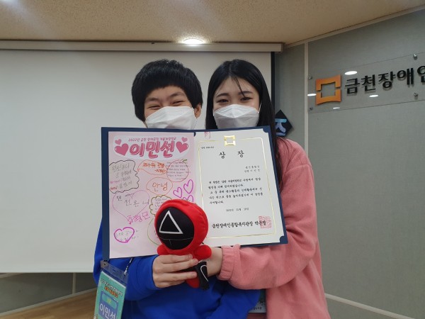 상장과 선물 받은 오징어 게임 인형을 들고 있는 참여자와 복지사 선생님이 함께 카메라를 보며 웃고 있는 모습