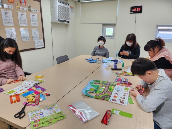 사각 테이블에 모여앉아 종이접기 책을 보고 색종이를 접는 단체사진