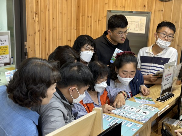 해오름 작은도서관에서 그림글자판으로 책 위치에 대해 문의하는 방법을 보여주는 AAC모니터링단원과 단원을 옹기종기 모여서 바라보는 참여자들의 모습