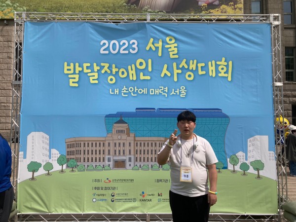 2023년 서울 발달장애인 사생대회 포스터 앞에서 포즈를 취하는 이용자