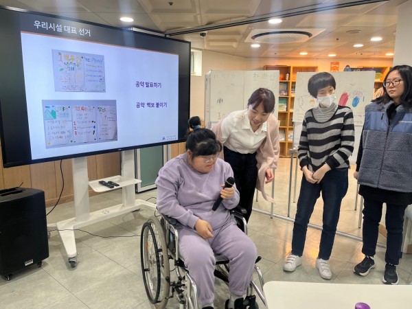 선거 공약 발표가 어려워 담당 사회복지사의 도움을 받아 발표를 이어가는 이용인분의 모습